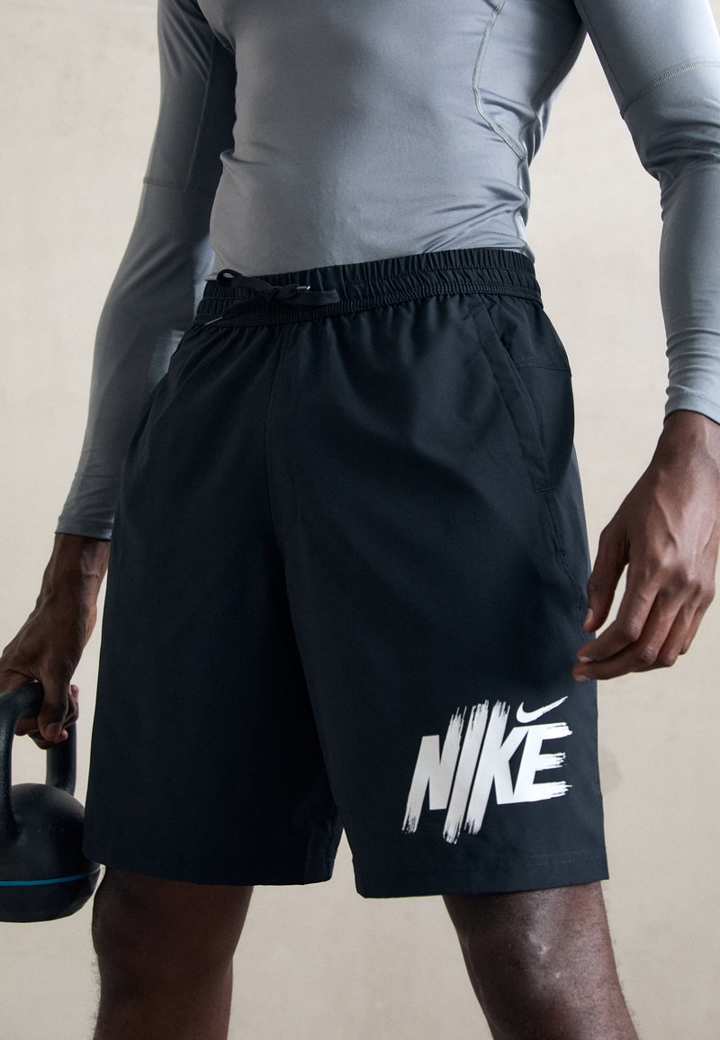 Vyras, vilkintis juodus Nike šortus ir pilką ilgomis rankovėmis marškinį, laiko juodą kettlebell'ą su mėlynu dryžiu, stovėdamas prieš paprastą foną.