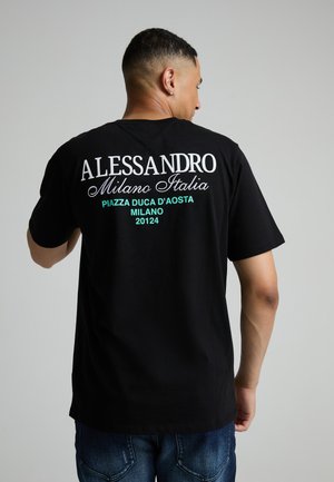 Mand iført sort T-shirt med hvid og grøn tekst, der refererer til Alessandro Milano Italia og Piazza Duca d'Aosta Milan 20124, set bagfra.