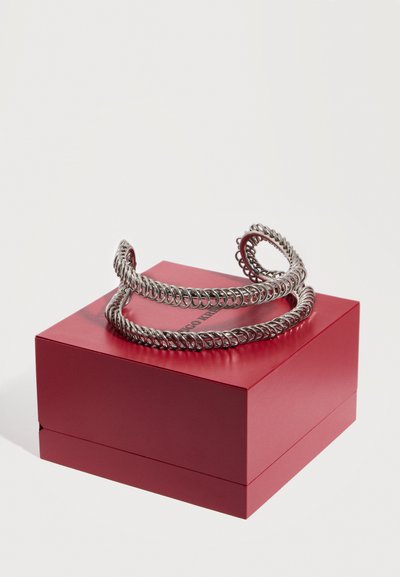 Ασημένιο βραχιόλι σε σχήμα σπείρας με στριφτή σχεδίαση, τοποθετημένο πάνω σε ένα κλειστό κόκκινο ορθογώνιο κουτί κοσμημάτων, σε λευκό φόντο.