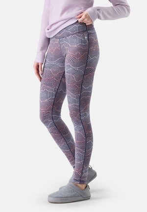Persona con leggings morados y rosas estampados con un pequeño bolsillo lateral con cremallera, camiseta lila clara y zapatos tipo slip-on grises.