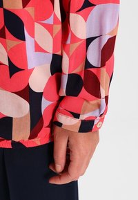 Gros plan sur la main d'une personne près d'une manche avec un motif géométrique coloré en rouge, rose, beige, noir et violet.