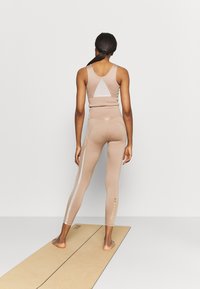 Senza cuciture reggiseno sportivo color nude con taglio a triangolo e leggings abbinati caratterizzati da una striscia bianca e dettagli dorati, posizionati su un tappetino per yoga.