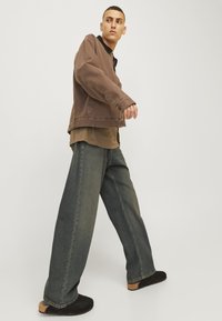 Young man walking, wearing a brown jacket, olive green shirt, wide-leg faded jeans, and black slip-on shoes against a plain light background.