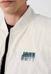 Witte bomberjack met een ritssluiting aan de voorkant, geribbelde kraag en geborduurd "JOTT" logo in schaduwen van blauw en grijs op het linkerborstgebied.