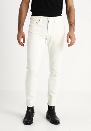 Homme portant un jean blanc slim, des bottines noires et une chemise noire rentrée dans le pantalon, debout sur un fond blanc uni.