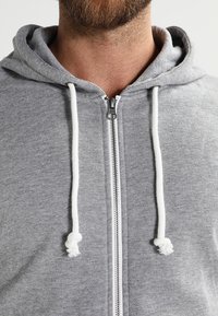 Grå zip-hoodie med vit dragsko, tillverkad av mjukt tyg. Har en standard framdragkedja och en design med känguruficka.