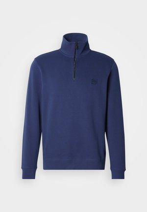 Marineblå genser med kvartglidelås i halsen, lange ermer og myk tekstur. Har en liten svart logopatch på brystet.