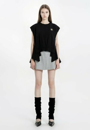 Joven con el pelo largo que lleva una camiseta sin mangas negra, minifalda gris, calcetines negros caídos y tacones negros con punta afilada, de pie mirando al frente.