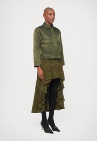 Olive rohelise satiinjakiga, millel on kaks rinna taskut, koos tumeda lillemustriga asümmeetrilise seelikuga, millel on volangid, mustad sukkpüksid ja kontsadega kingad.