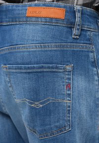 Vaqueros de mezclilla en un tono azul medio, con parche de cuero de la marca y costuras decorativas en los bolsillos traseros. Tela texturizada, corte clásico.