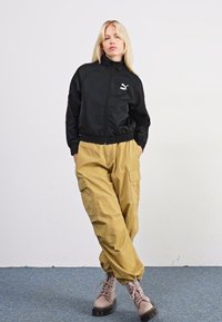 Schwarze, leichte Zip-Jacke mit hohem Kragen und Puma-Logo, kombiniert mit beigefarbenen Cargo-Hosen mit Taschen. Hellbraune Schnürstiefel.