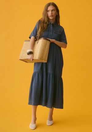 Vrouw in een denim midi-jurk die een grote gevlochten mandtas vasthoudt, witte gevlochten hakken draagt en twee armbanden op een gele achtergrond.
