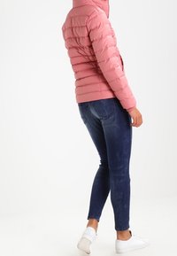 Rosa vadderad jacka med horisontell quiltning och figurnära design, kombinerad med mörkblå jeans och vita sneakers.