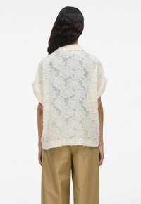 Blusa de encaje en blanco roto con patrón floral, espalda transparente, mangas cortas y dobladillo deshilachado; combinada con pantalones anchos de un beige claro.