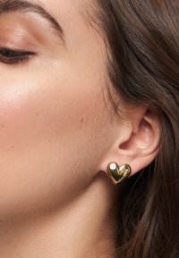 Goldenes, herzförmiges Ohrstecker, mit glatter Textur und glänzender Oberfläche, auffällig am Ohr präsentiert, wobei die abgerundeten Kanten und die reflektierende Fläche hervorgehoben werden.