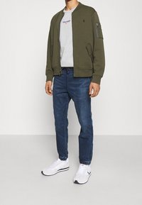 Man med olivfärgad bomberjacka, grå grafisk t-shirt, blå jogger-stil jeans och vita sneakers står mot en vit bakgrund.