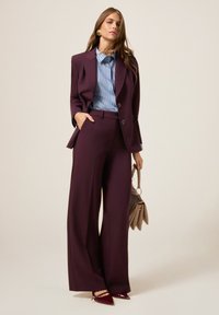 Bordový oblek s širokými kalhotami, sakem na míru a modrou pruhovanou košilí. Doplňky zahrnují béžovou tašku a burgundské lodičky.