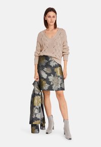 Beige Strickpullover mit V-Ausschnitt, kombiniert mit einem figurbetonten schwarzen Rock mit gold- und silberfarbenen Blumenmustern. Graue Knöchelstiefel runden das Outfit ab.