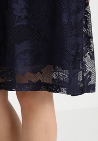Jupe en dentelle bleu marine foncé à motif floral, partiellement transparente, portée au-dessus de jambes nues sur un fond clair.