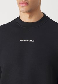 Musta pehmeästä kankaasta valmistettu collegepaita, jossa on pyöreä kaula-aukko ja hillitty valkoinen "EMPORIO ARMANI" -logo keskellä rintaa.
