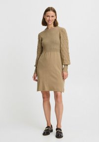 Stickad klänning i beige med ribbat liv och texturerade, mönstrade ballongärmar. Kanten slutar strax ovanför knäet. Matchas med svarta loafers.
