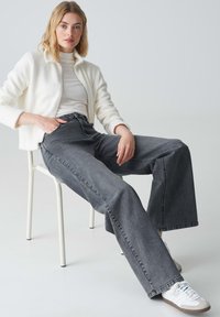 Hoch taillierte, weit geschnittene graue Jeans aus Denim, kombiniert mit einem eng anliegenden weißen Rollkragenpullover und einer weichen, cremefarbenen Zip-Jacke, getragen mit weißen Sneakern.
