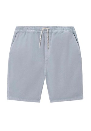 Homme debout, de dos, portant une casquette verte, un t-shirt bleu clair oversize, un short vert menthe et des baskets blanches sur fond beige.