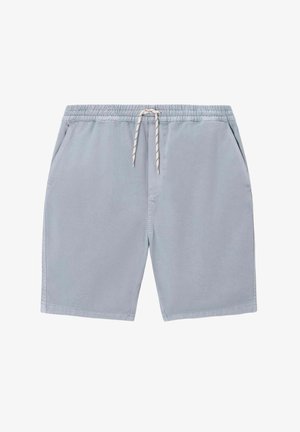 Homme debout, de dos, portant une casquette verte, un t-shirt bleu clair oversize, un short vert menthe et des baskets blanches sur fond beige.