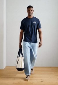 Tamsiai mėlynas medvilninis marškinėliai su logotipu ant krūtinės, derinami su šviesiai mėlynomis džinsomis. Nešiojama kreminė ir tamsiai mėlyna drobės pirkinių maišelis.