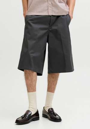 Mężczyzna w luźnych czarnych spodenkach do kolan, beżowych skarpetkach, ciemnobrązowych mokasynach z frędzlami oraz w kraciastej koszuli zapinanej na guziki, z rękami w kieszeniach.