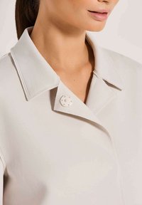 Frau in einem hellen Mantel mit einem weiten Kragen und einem dekorativen runden Knopf in der Nähe des Ausschnitts.