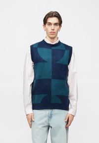 Πλεκτό γιλέκο σε σκούρο μπλε και τιρκουάζ με γεωμετρικά σχέδια. Αμάνικο, στρογγυλή λαιμόκοψη, φορεμένο πάνω από ανοιχτόχρωμο πουκάμισο, υφασμάτινη υφή.