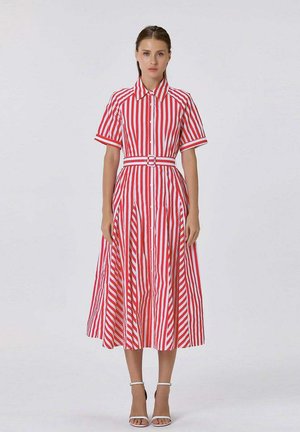 Femme debout portant une robe midi rayée verticale rouge et blanche avec manches courtes, col, ceinture, et sandales blanches à talons hauts.