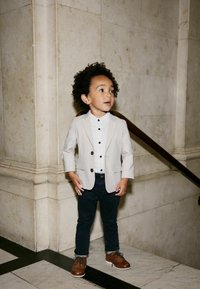 Enfant portant un blazer gris clair, une chemise blanche à boutons noirs, un pantalon bleu marine et des chaussures marron, se tenant devant un mur en marbre avec un escalier.