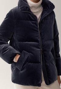 Osoba ubrana w granatową welwetową pikowaną kurtkę na beżowym golfie, z jedną ręką w kieszeni kurtki.