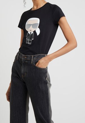 Kobieta w czarnej koszulce z nadrukiem postaci w okularach przeciwsłonecznych oraz w ciemnych, wysokich dżinsach z jedną ręką w kieszeni.