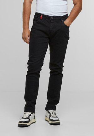 Mann, der schwarze Slim-Fit-Jeans, weiße und schwarze Sneakers und ein weißes Tanktop trägt, mit einer Hand in der Tasche.