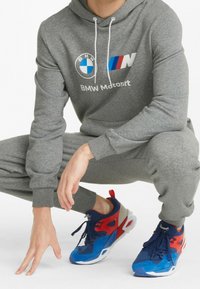 Grijze hoodie met BMW-logo en tekst, gecombineerd met sneakers in kleurblokken in blauw, rood en grijs. Sportieve stof, relaxte pasvorm.