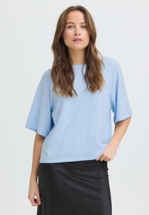 Vrouw met lang bruin haar, gekleed in een lichtblauw kortemouwen top en een zwarte satijnen rok, staand tegen een effen lichte achtergrond.