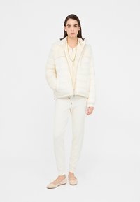 Giacca puffer color crema, con zip frontale, cappuccio e cuciture orizzontali. Abbinata a pantaloni jogger coordinati in crema e scarpe flat chiare.