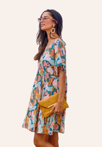 Robe florale colorée à manches courtes, avec un décolleté en V et une jupe à volants. Complétée par une pochette jaune et des boucles d'oreilles surdimensionnées.