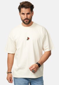 Cremfarbendes, übergroßes T-Shirt aus Baumwolle mit einem kleinen, bunten gestickten Design auf der Brust. Kurze Ärmel und runder Halsausschnitt.