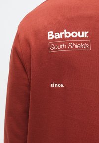 Κοντινό πλάνο μιας μπλούζας σε καστανοκόκκινο χρώμα με λευκή επιγραφή που γράφει "Barbour South Shields" και "since." στην περιοχή του ώμου στο πίσω μέρος.