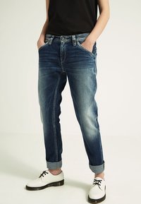 Jeans i denim i mörkblått med urtvättade områden och uppvikt fåll; har fem fickor och knappgylf. Matchas med vita skor.