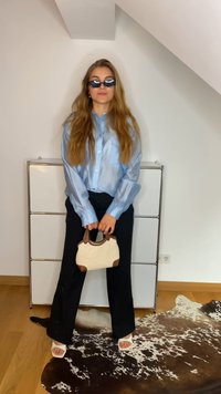 Camisa azul clara transparente com botões, calças largas pretas, sandálias brancas, segurando uma mala bege com detalhes castanhos, sobre um tapete padrão.