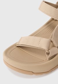 Beige sandal featuring a textured rubber sole, adjustable fabric straps, and a flat design, ideal for comfort and ease of wear.