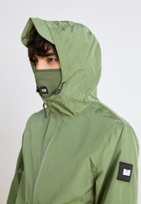 Lightweight, olive green jacket with a high collar and hood, featuring a matching face mask. Includes a fabric badge on the left sleeve.