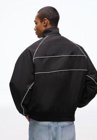 Black bomber jacket with white piping details, elastic waistband, and a high collar. Made from a smooth, lightweight material.