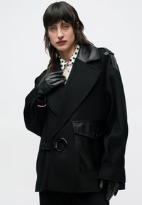 Čierny voľný kabát s vonkajšou stranou z vlny a koženými detailmi. Má veľkú prednú kapsu, opasok v páse a špicatý golier.