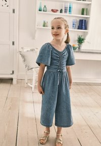 Denim jumpsuit med korte puffærmer, rynket bærestykke, indsnøret talje og brede cropped bukser. Parret med gyldne sandaler med stropper.